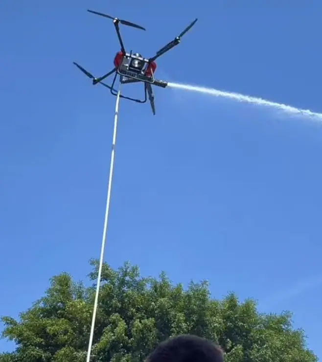 Un drone relié au sol par un tuyau pulvérise en plein ciel au-dessus des arbres.